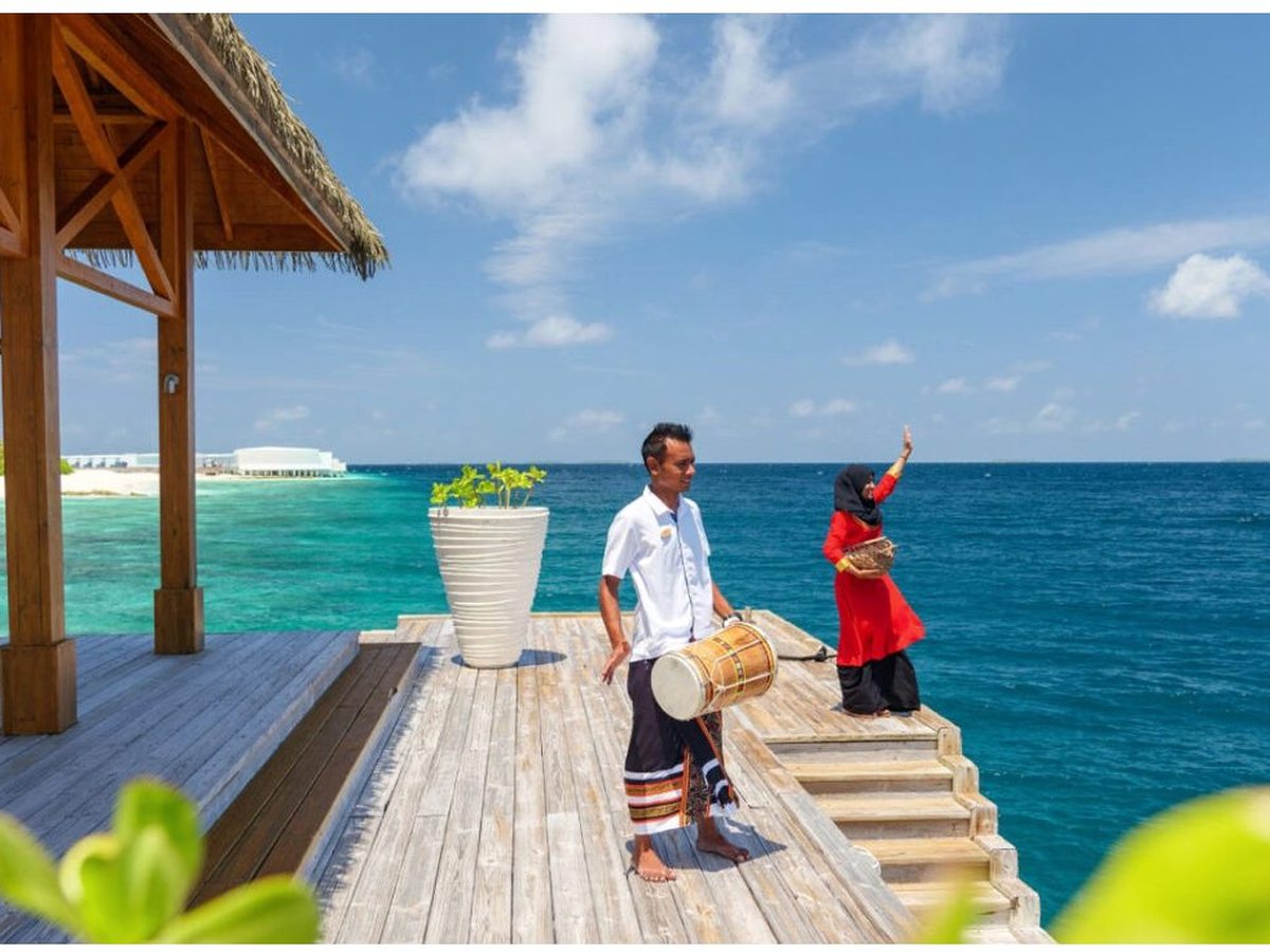 Two Amilla staff in traditional dress on a timber deck over turquoise water, one holding a Maldivian drum and one waving, with a thatched pavilion to the side.