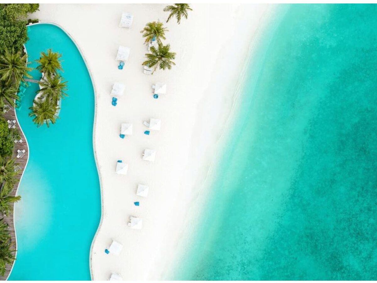 A large freeform freshwater pool edged with cabana daybeds, set on a white sand beach beside a turquoise lagoon, seen from above.