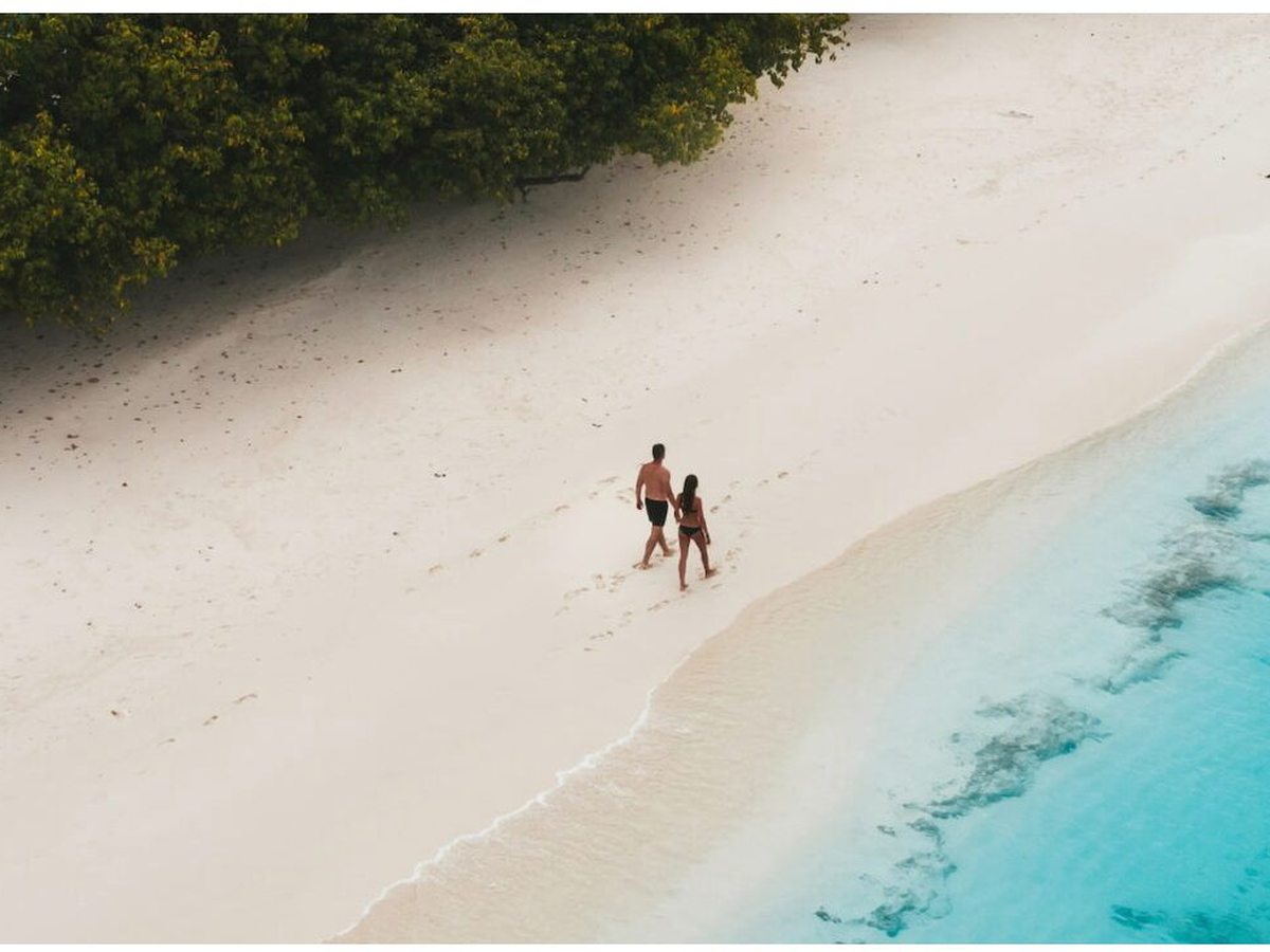 A wide, near-empty white sand beach curving along the turquoise lagoon, with two people walking at the water's edge.