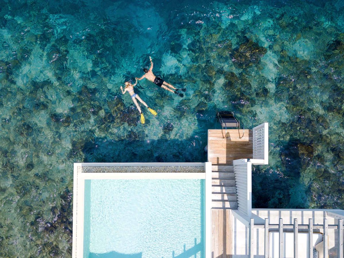 An overwater villa's plunge pool and timber swim deck seen from above, beside clear water and coral reef where two people snorkel.
