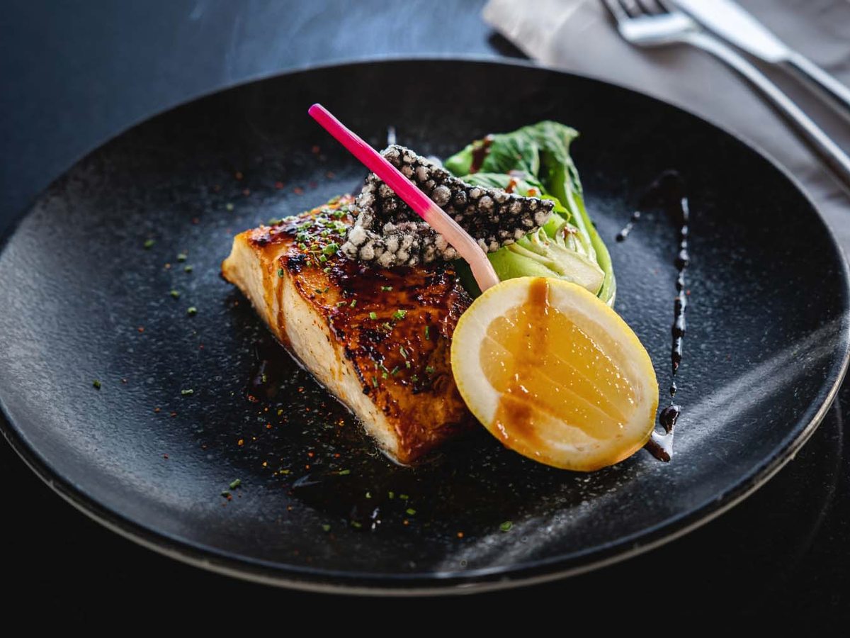 A fillet of fish plated on a dark round plate with greens and a wedge of lemon.