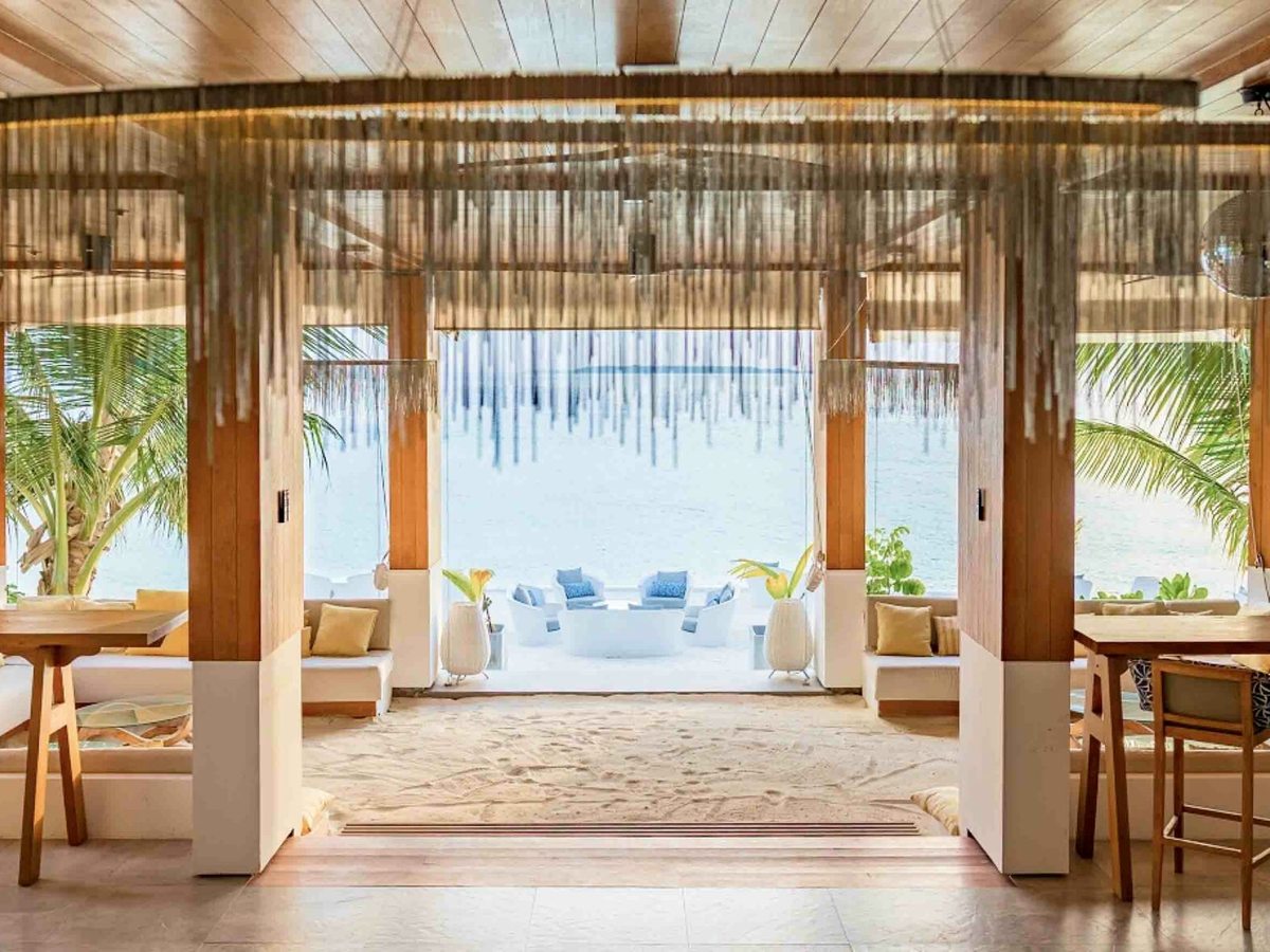 An open-sided beachfront dining room with timber tables and chairs, hanging fringe screens and a sand floor, looking out to palms and the lagoon.