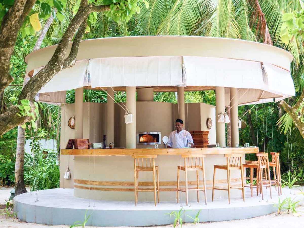 A circular open-air beach bar with a pale canopy roof and a curved timber counter with stools, set on the sand among palms.