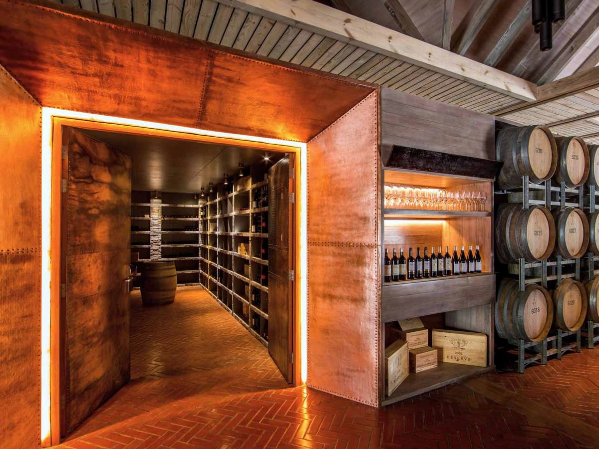 A warmly lit wine cellar with an arched timber entrance, bottle racks along the walls and wooden barrels stacked at the far end.