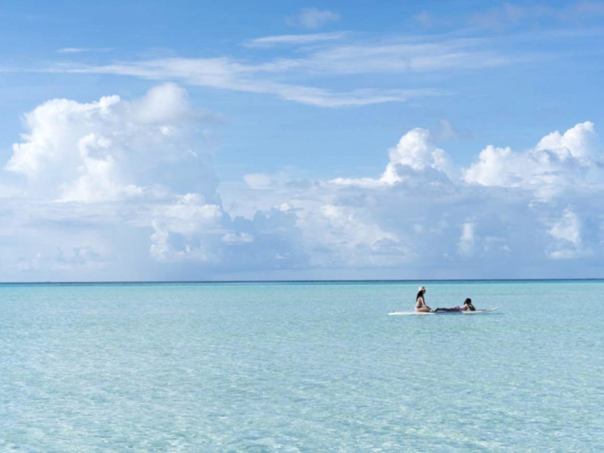 Two people on a stand-up paddleboard far out on the calm, clear turquoise lagoon under a bright sky with scattered clouds.