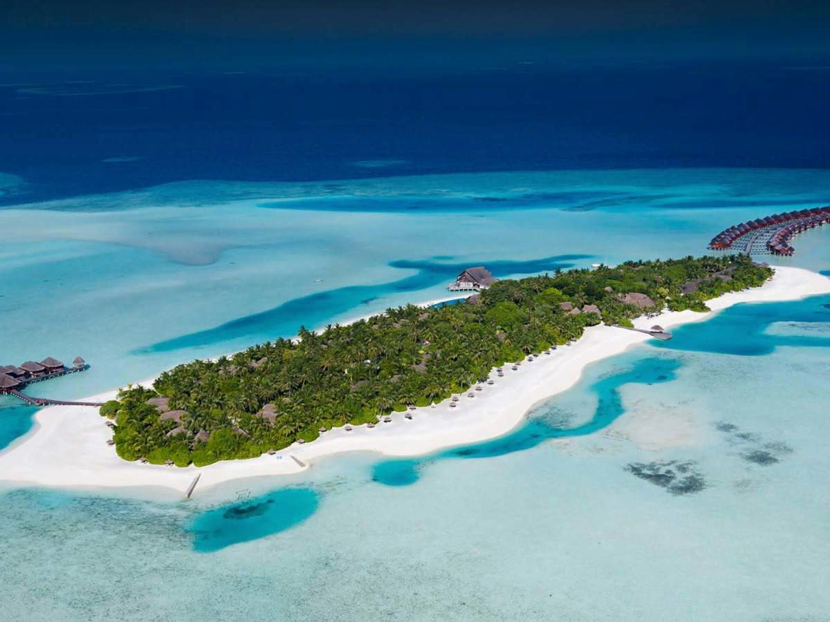 Aerial view of the resort island fringed with white sand and overwater villas, set in a broad turquoise lagoon with a sandbar.