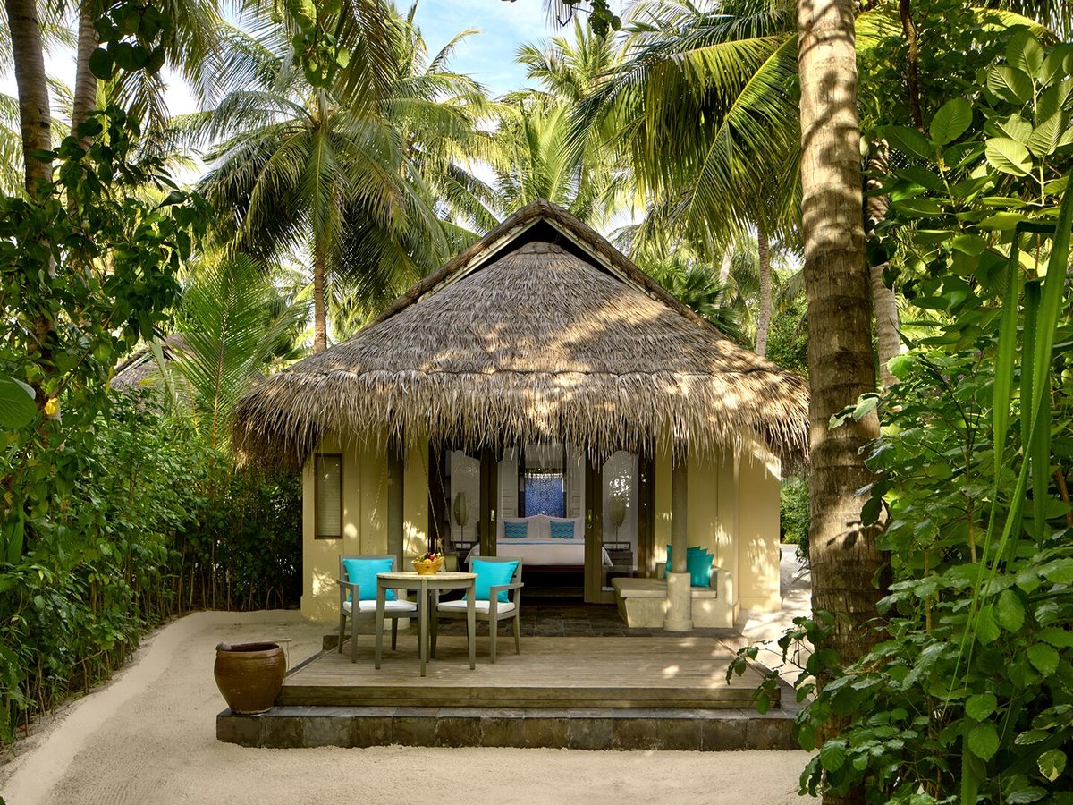 A thatched beach villa with a covered terrace, table and chairs, set among tall palms at the end of a raked sand path.