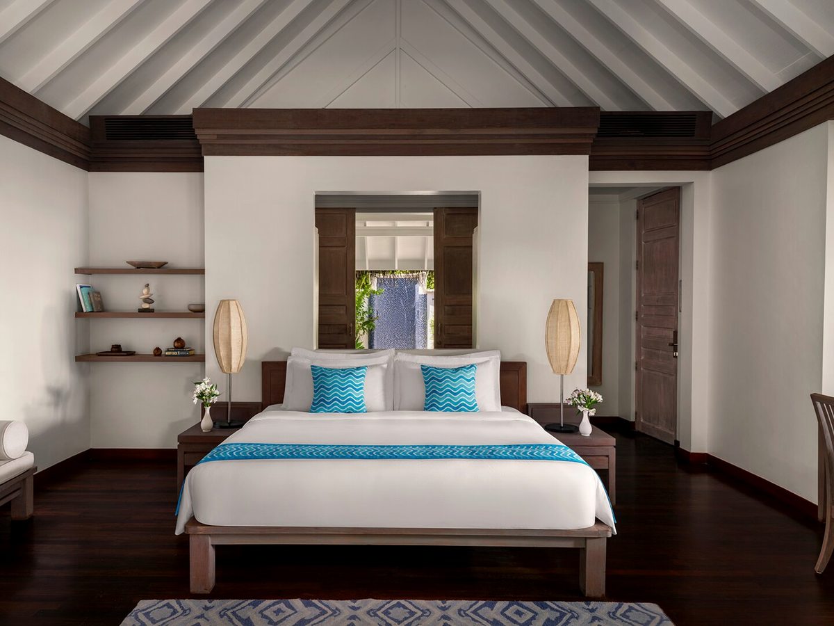 A villa bedroom with a king bed, turquoise runner and cushions, a writing desk and windows onto tropical greenery under a vaulted timber ceiling.