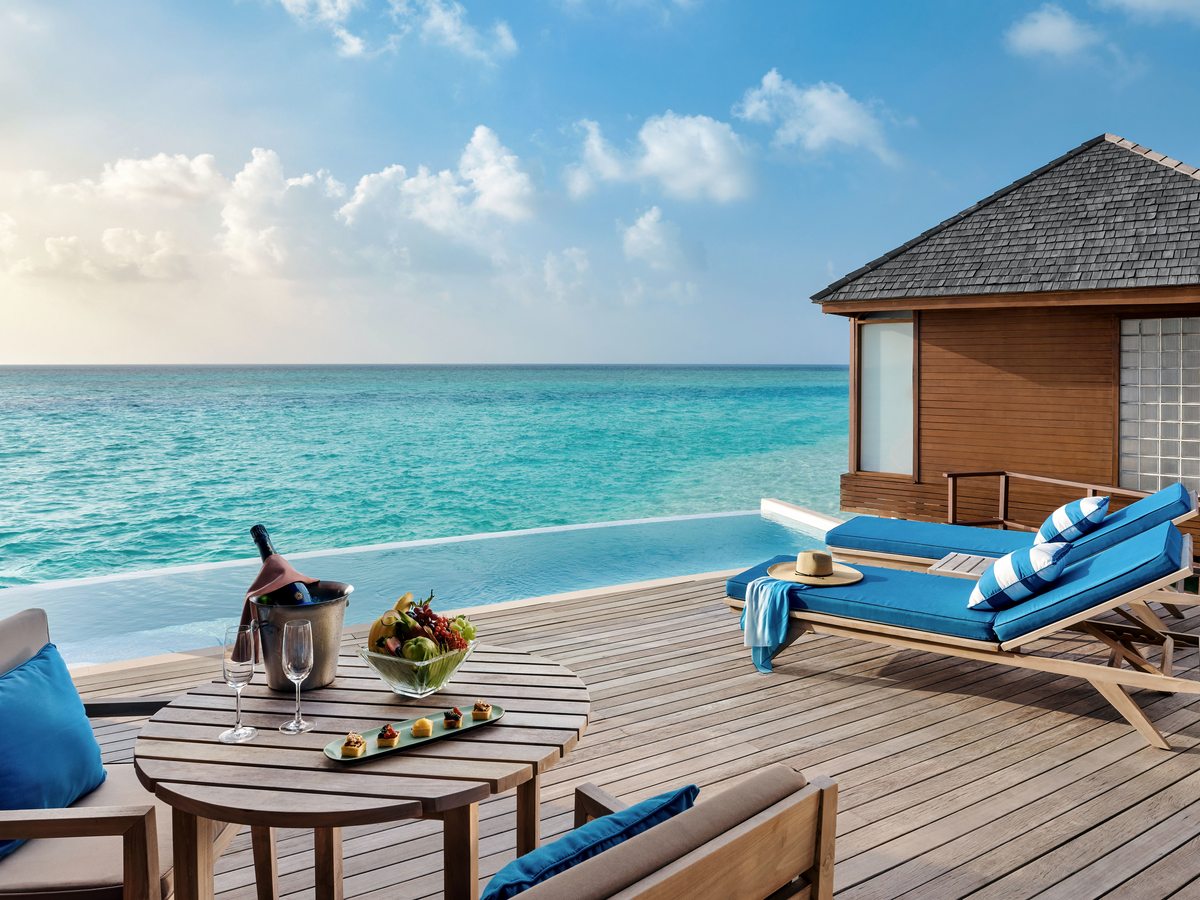 A private infinity pool on an overwater villa's timber deck, with blue-cushioned loungers and an outdoor table, above a turquoise sea.