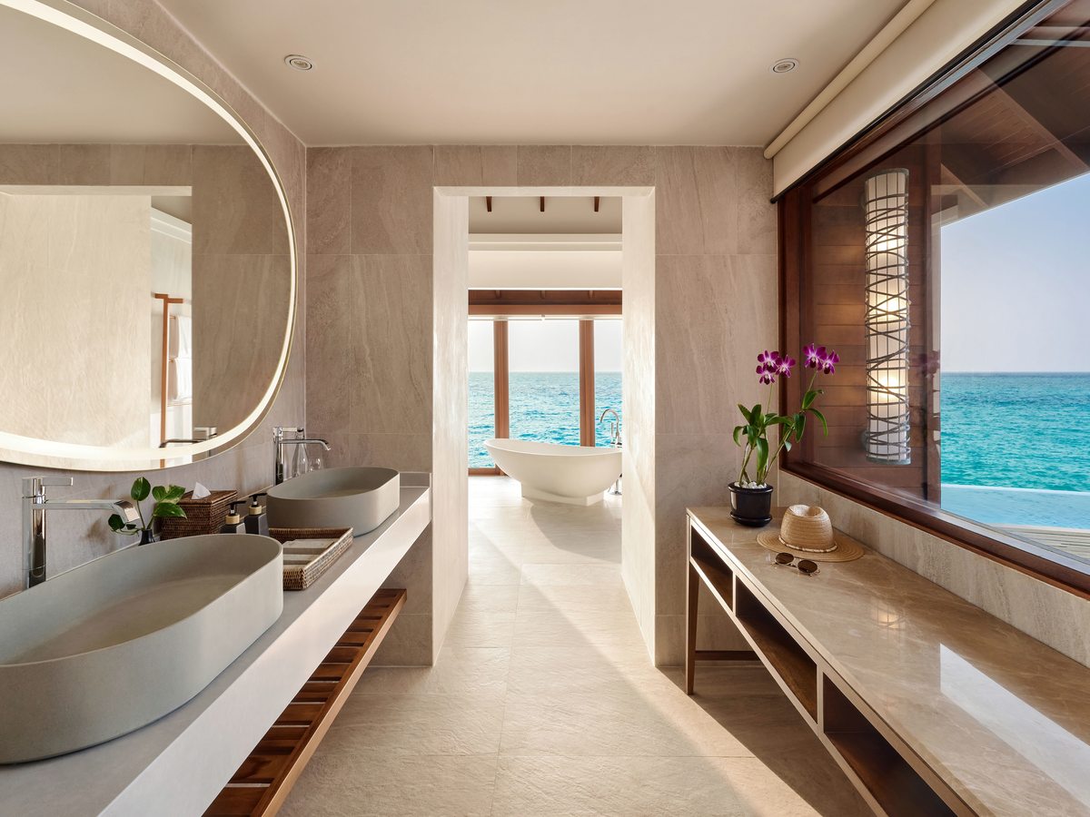 An open-plan villa bathroom with twin basins on a stone counter, an illuminated mirror and a freestanding bath beside full-height windows onto the lagoon.