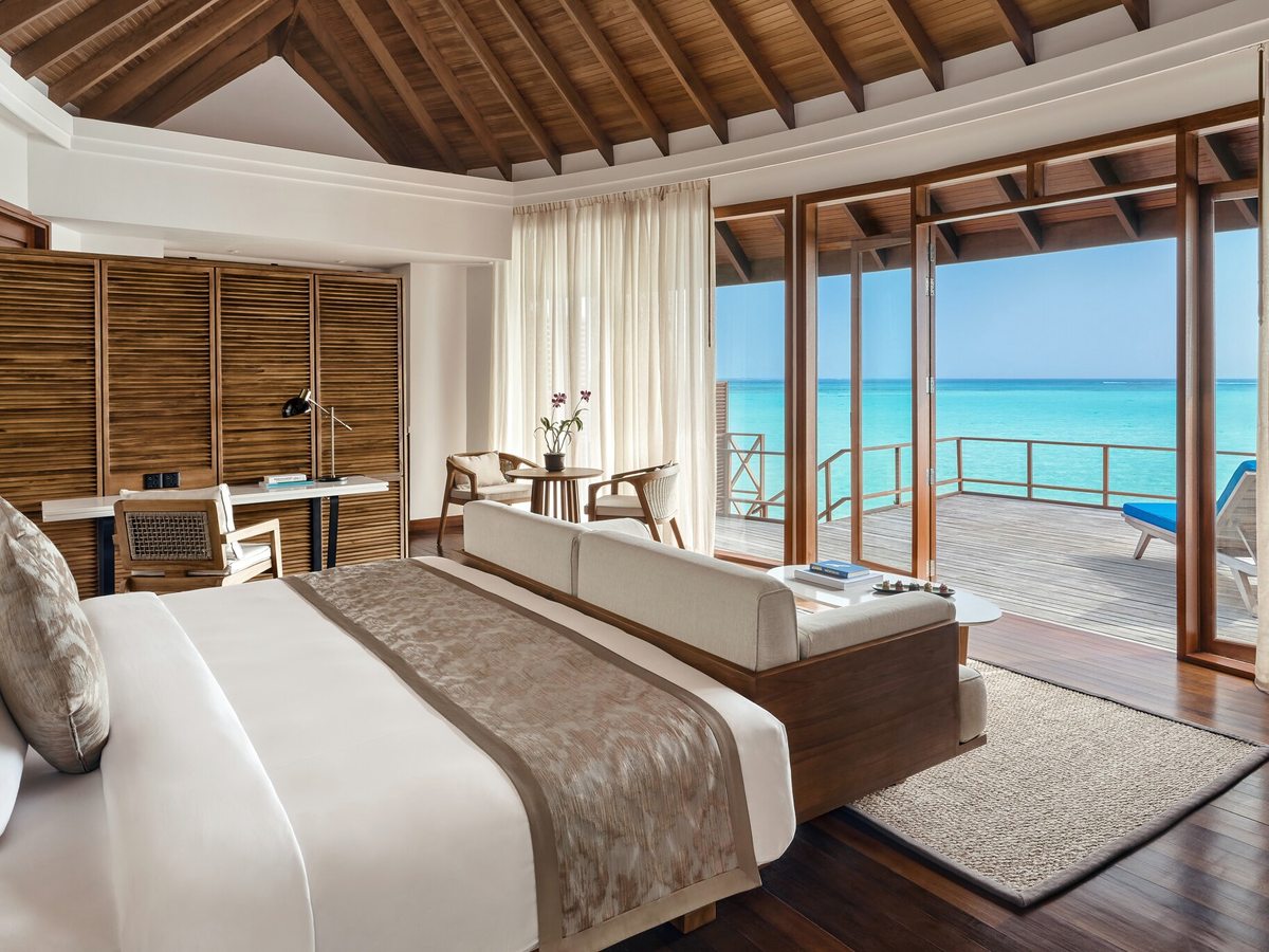 A bedroom with a king bed facing floor-to-ceiling glass doors that open to a private deck with sun loungers over a turquoise lagoon.