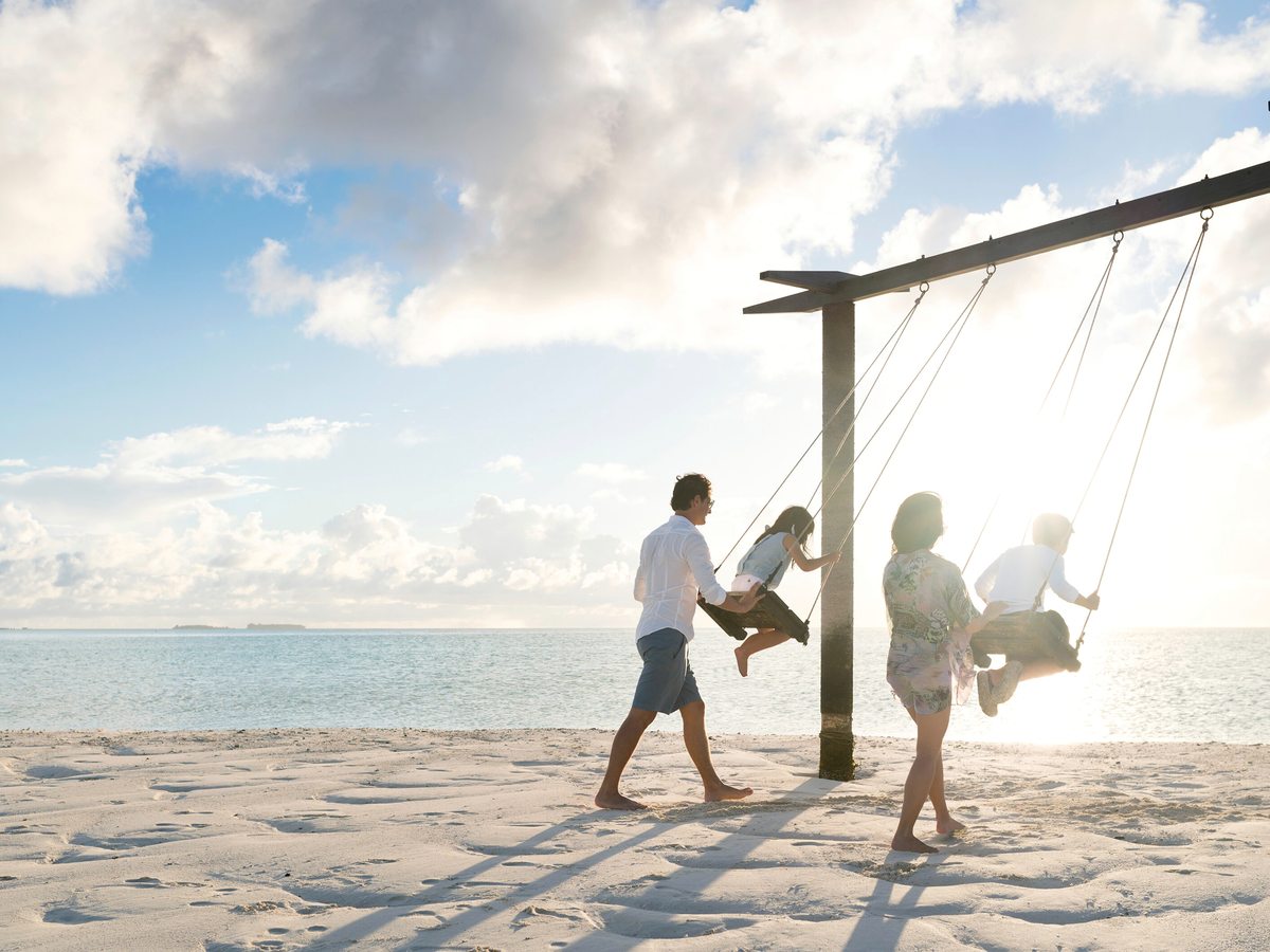 A parent and two children on a wooden swing on the sand at the water's edge, backlit by a bright sky over a calm sea.