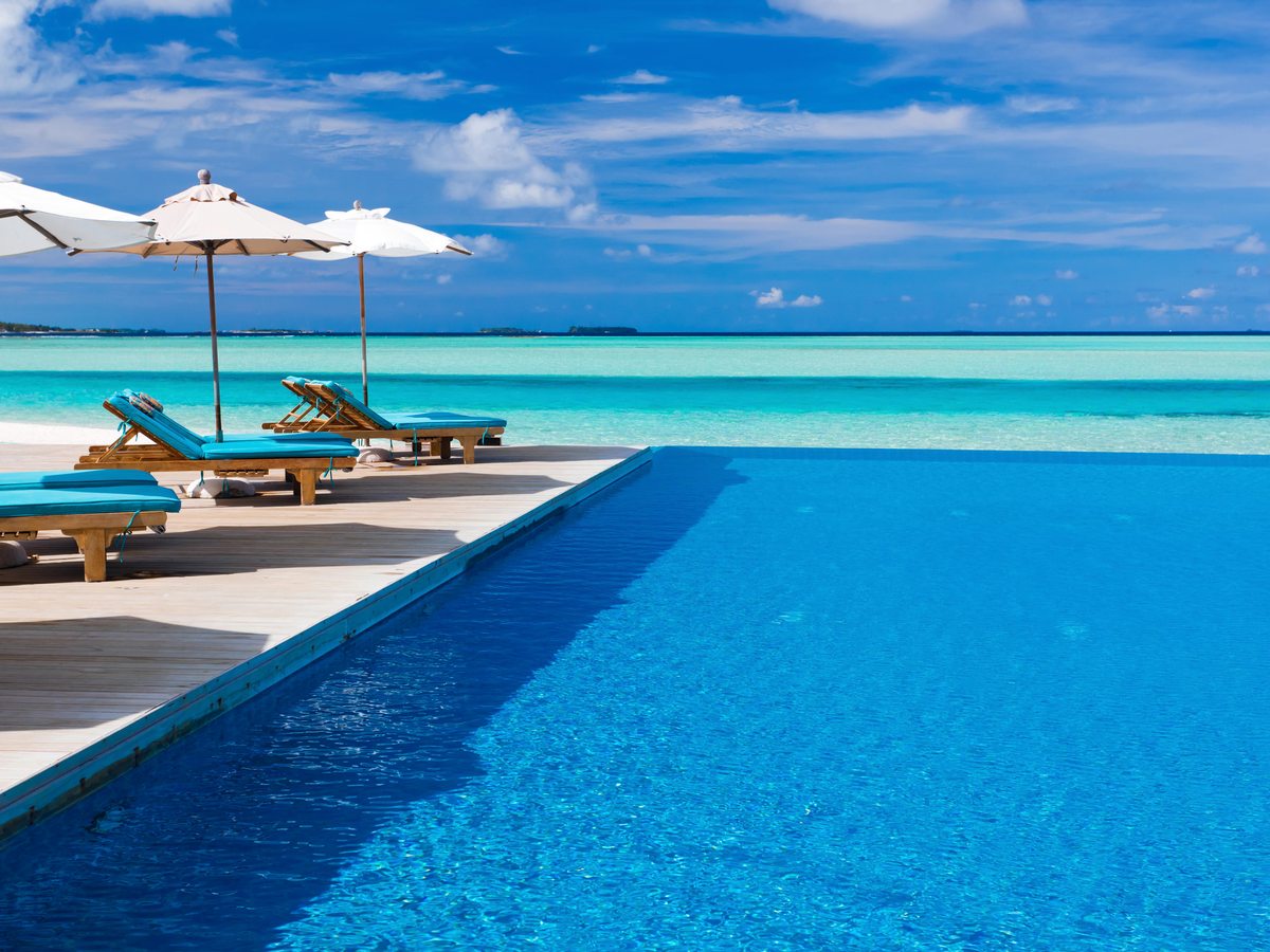 A large beachfront infinity pool edged with turquoise sun loungers and white parasols, looking out over the lagoon to the horizon.
