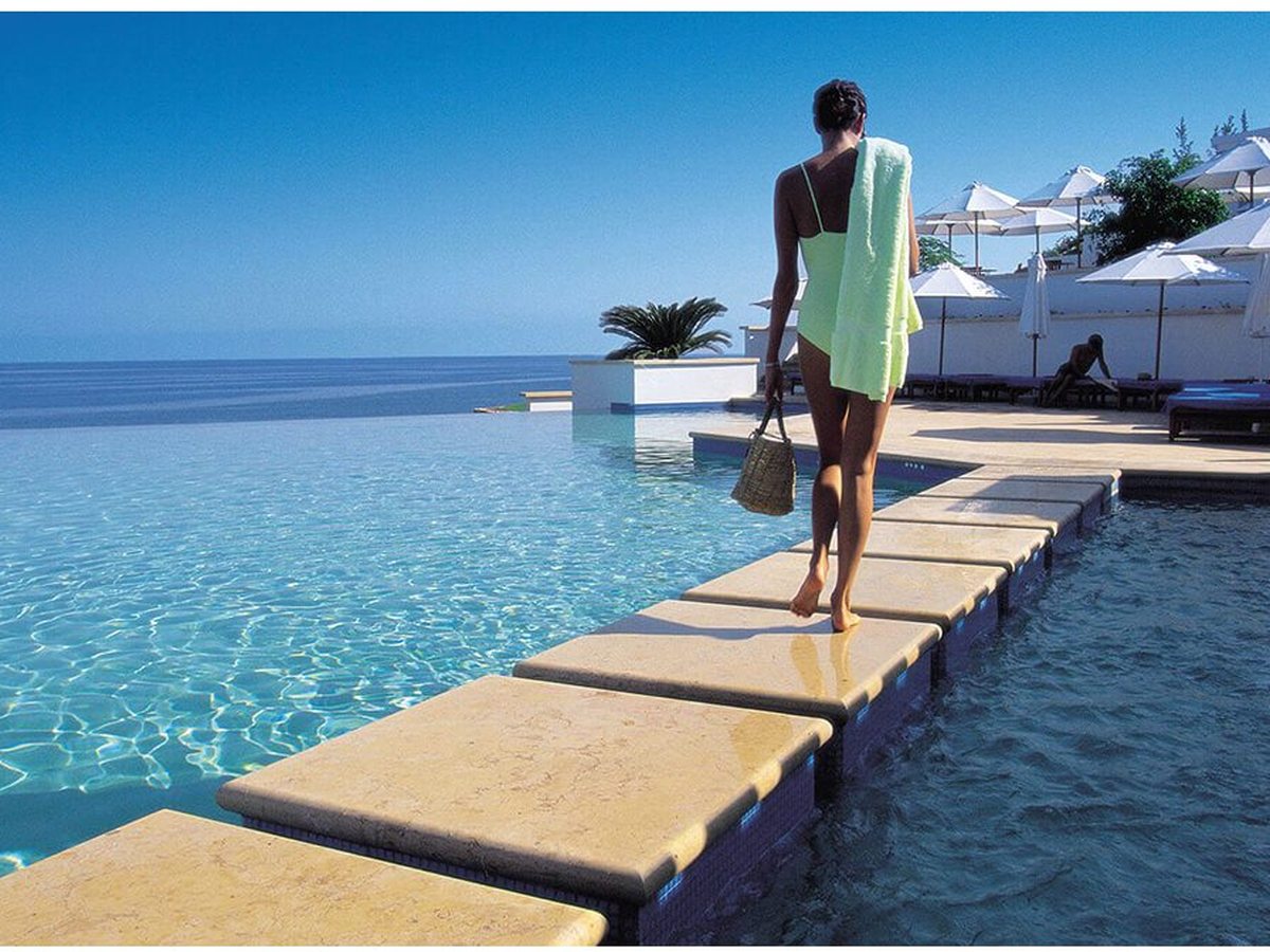 An infinity-edge pool with pale marble stepping stones across the water, a woman walking them with a towel, white parasols and the sea beyond.