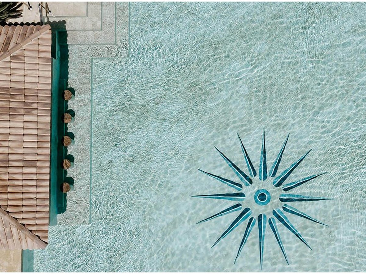 An overhead view of a pale-floored pool with a blue sunburst mosaic set into it, a tiled roof and the pool's green stone edge at the side.