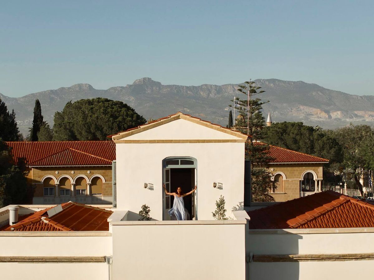 A white-walled rooftop terrace ringed by red-tiled roofs and sandstone arcaded buildings, a cypress and pine trees, mountains rising behind.