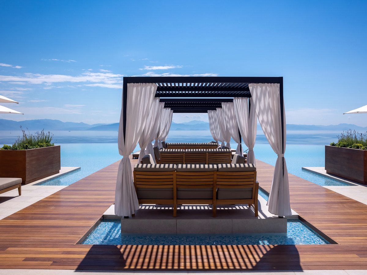 A four-poster cabana with white curtains and a daybed on the timber pool deck, framed against the sea and sky.