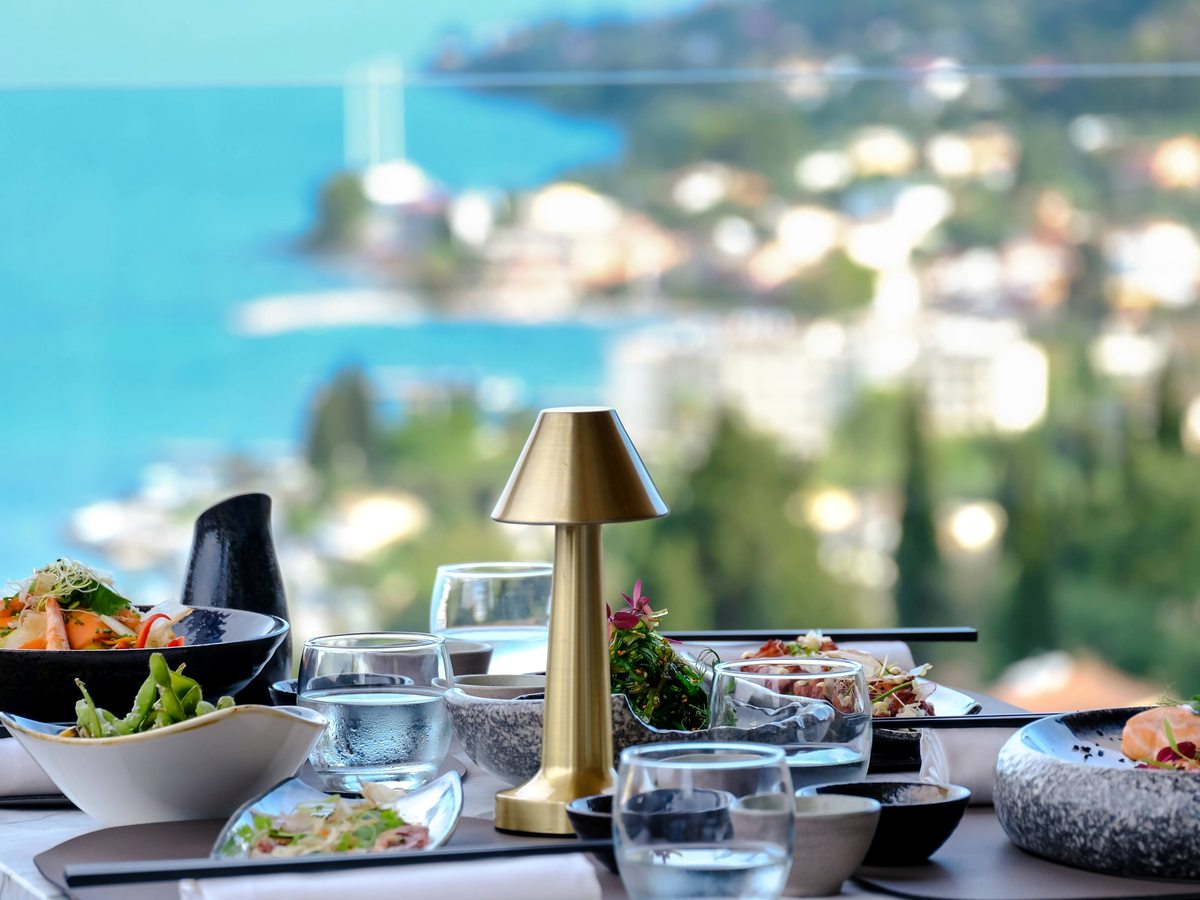 A table laid with Asian-style small plates, water glasses and a brass lamp, the blurred coast and bay of Benitses beyond.