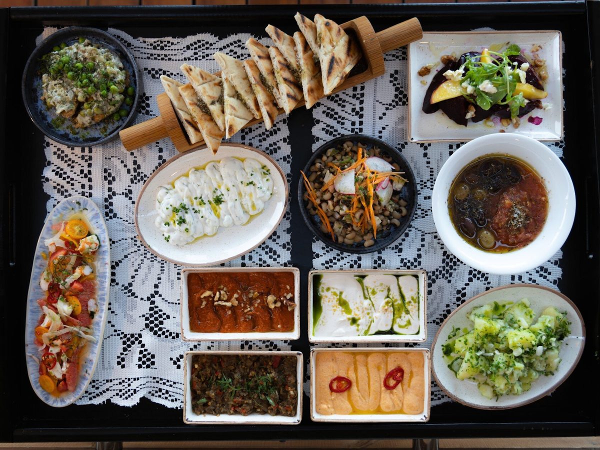 An overhead view of a spread of Greek mezze in small bowls and dishes, with flatbread and dips on a lace cloth.