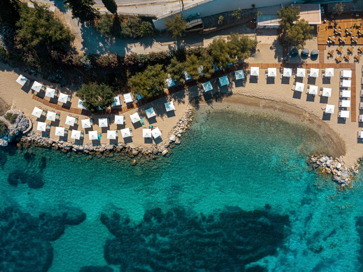 An aerial view of a small sandy cove lined with white parasols and sunbeds, clear turquoise water and rocks breaking the surface.
