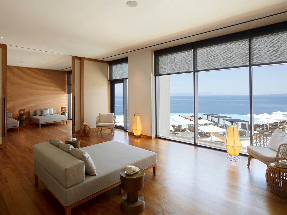 An open-plan suite living room with pale sofas and floor-to-ceiling windows looking over the pool loungers and the sea.