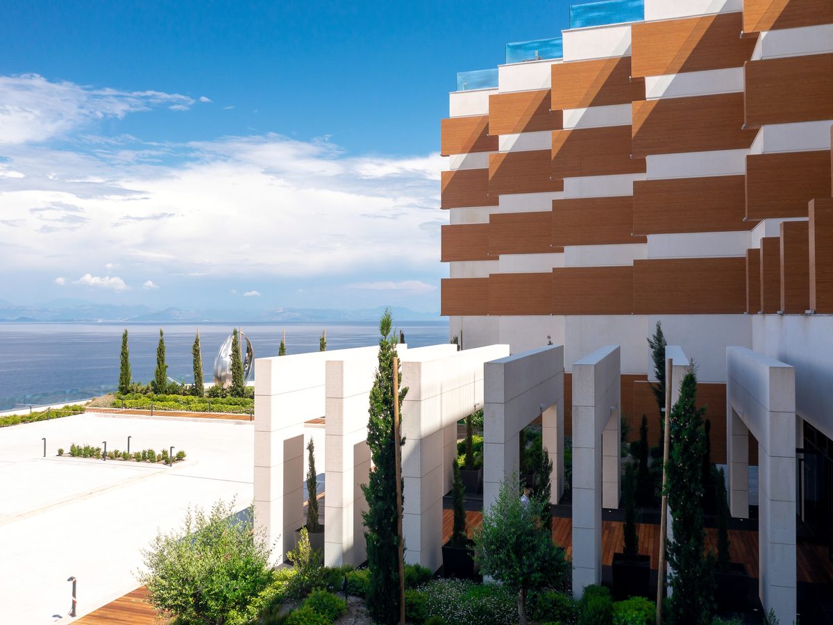 Modern white-and-timber resort buildings with terraced balconies stepping down a green hillside above the sea, cypress trees in the grounds.