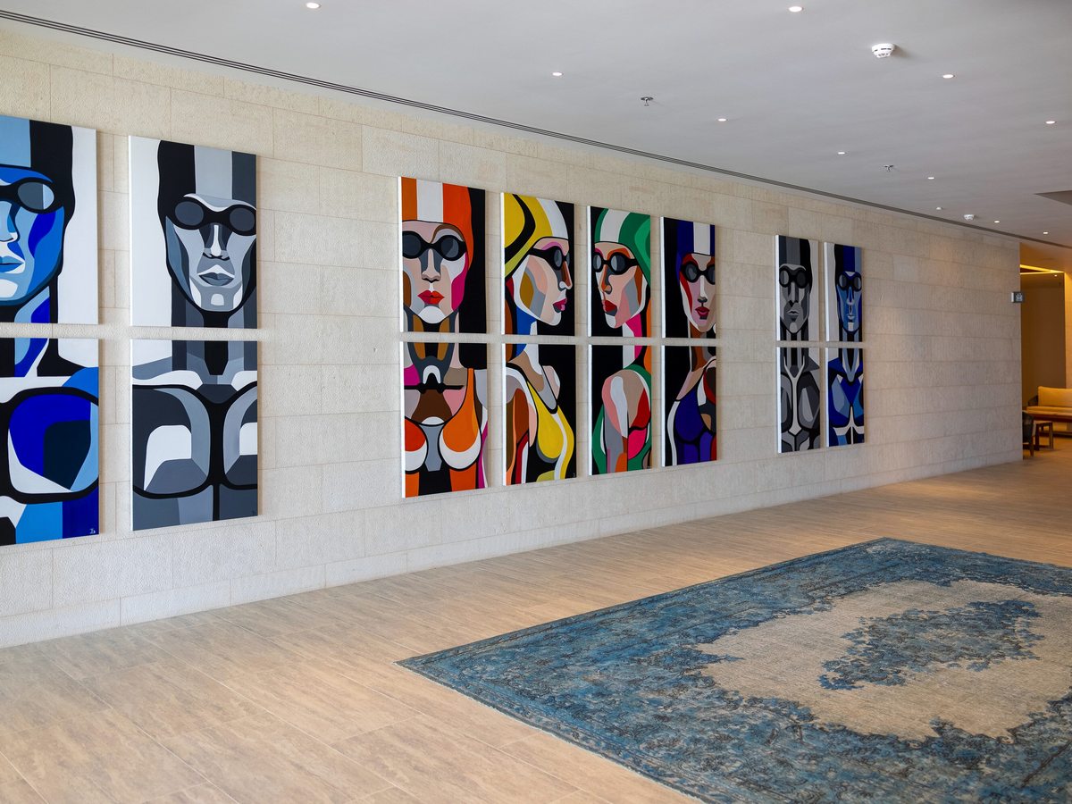 A long lobby corridor hung with a row of colourful pop-art portraits above a blue rug on a pale stone floor.