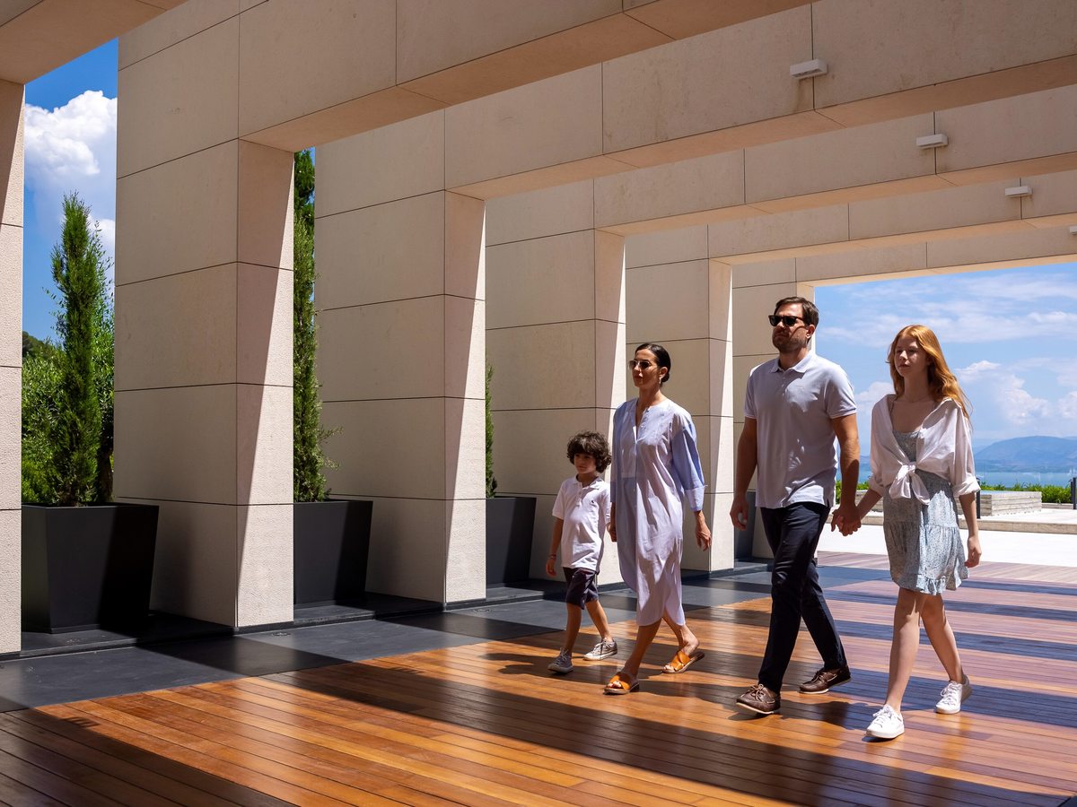 A family walking along a covered colonnade of tall cream columns on a timber floor, open to the sea on one side.