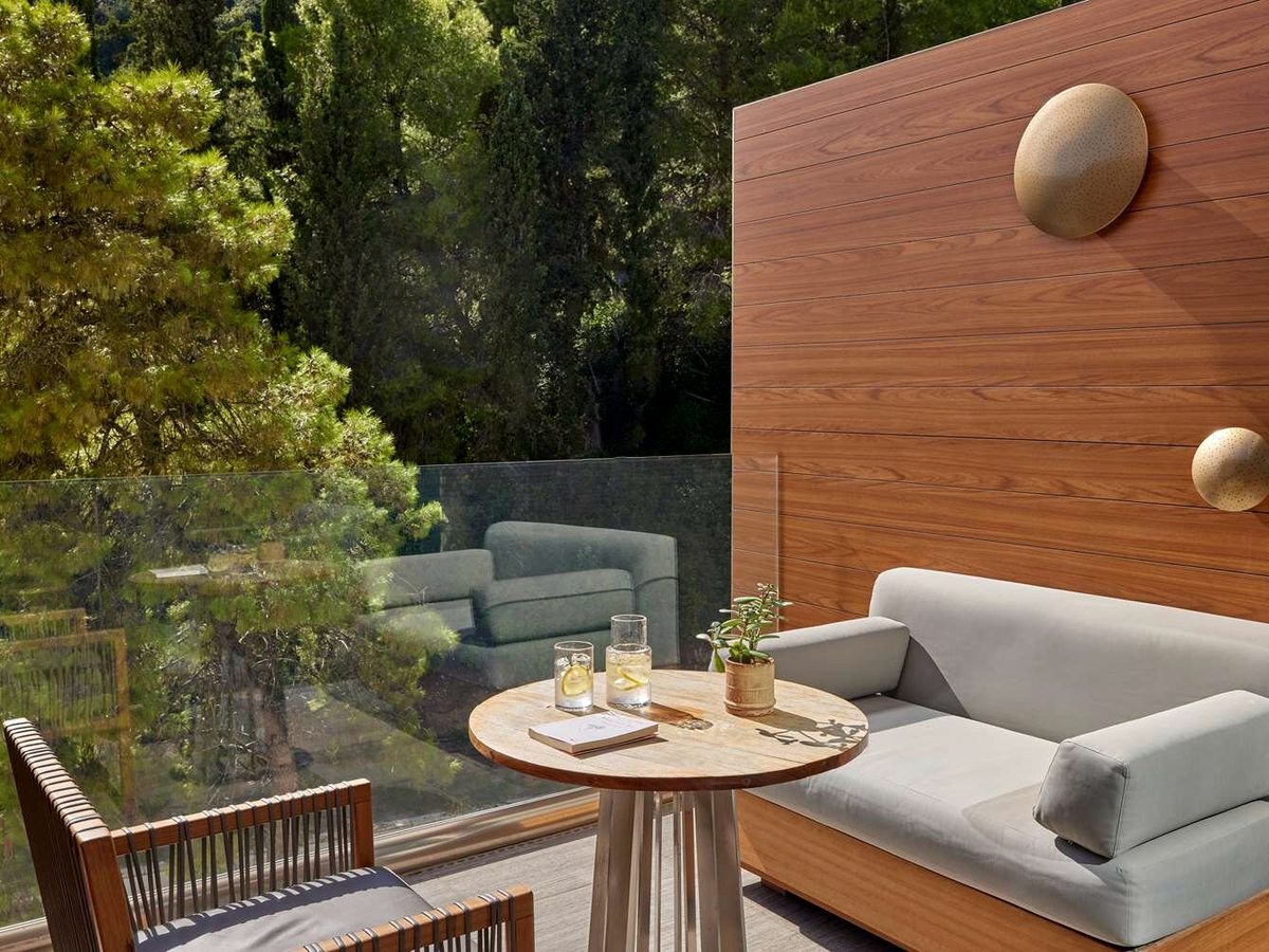 A private terrace with a built-in sofa and a small round table behind a glass balustrade, pine woodland rising directly behind.