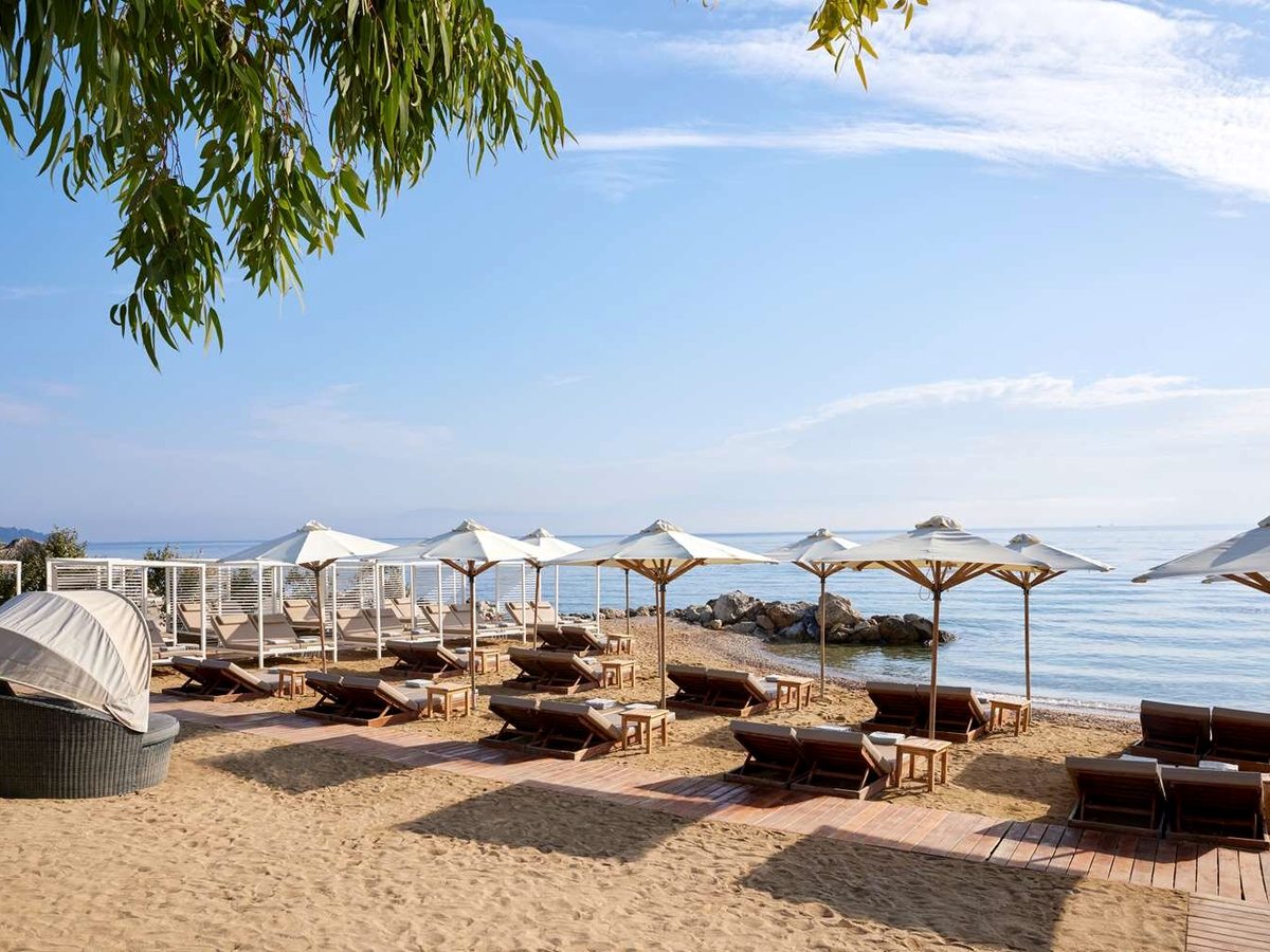 Rows of wooden sunbeds and white parasols on golden sand beneath an overhanging tree, a calm sea and a rocky point beyond.