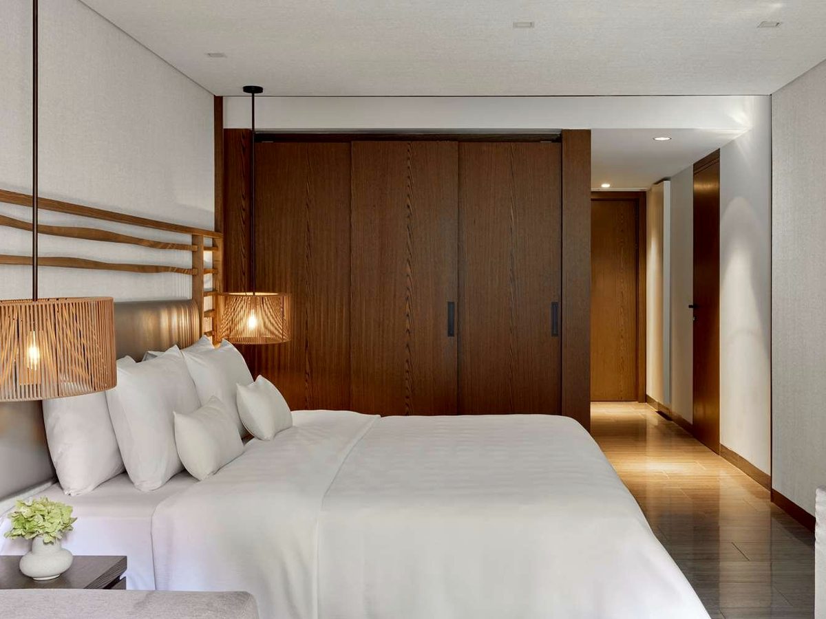 A bedroom with a king bed dressed in white, a timber-slat headboard, woven pendant lamps and a wood-panelled wardrobe wall leading to the entrance.