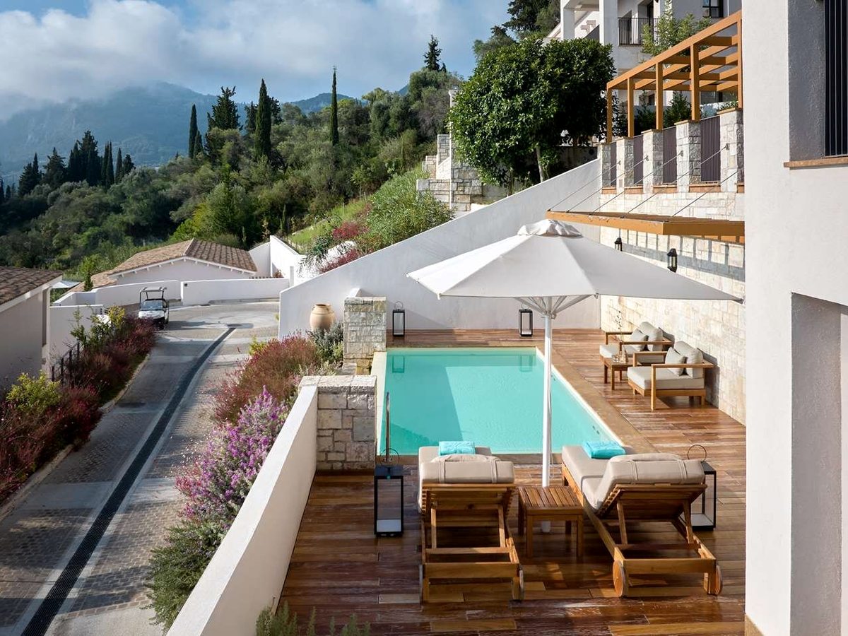A small private plunge pool with steps and a white parasol on a timber terrace, resort buildings and greenery rising on the hillside behind.