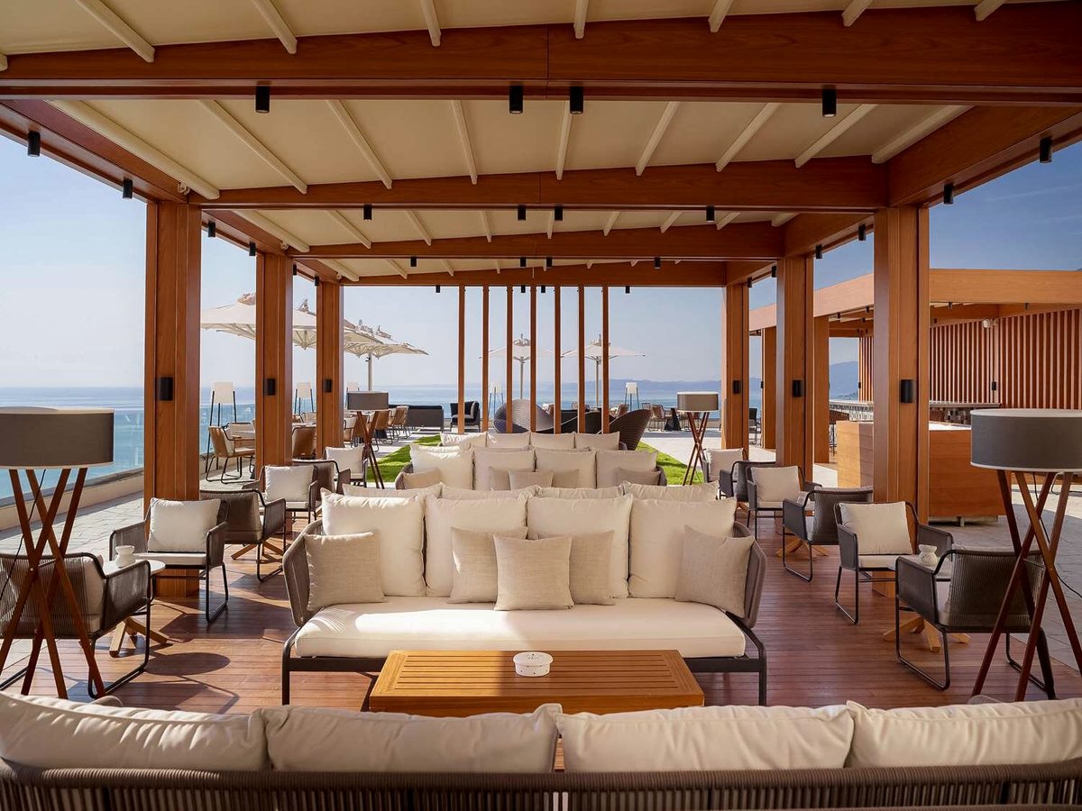 A covered lounge with cream sofas and armchairs under a timber pergola ceiling, opening to a sun terrace with parasols and the sea beyond.