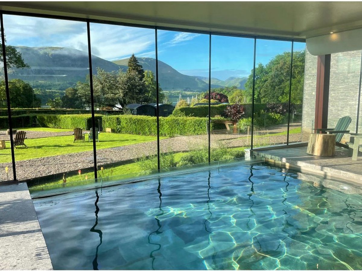 An indoor pool with sunlit turquoise water and a floor-to-ceiling glass wall looking out to a lawn, hedges and fells.