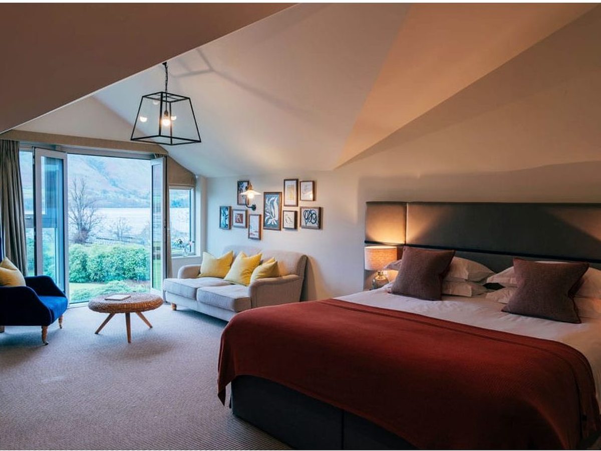 A spacious bedroom under a pitched ceiling with a red throw, a sofa-and-armchair sitting area and glazed doors to a garden with the lake beyond.