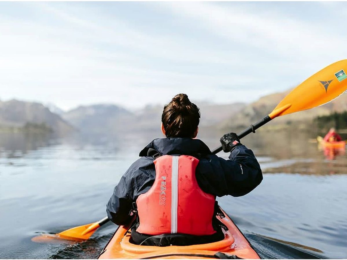 The view over the bow of an orange kayak, paddle raised, on the lake with fells ahead and another kayaker nearby.