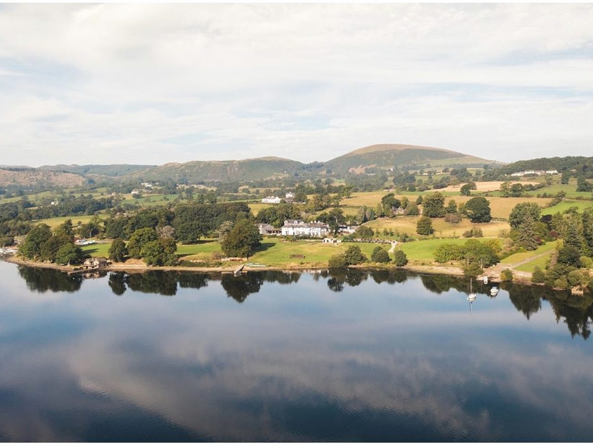 An aerial view over green fields and woodland to a still stretch of Ullswater and low fells.