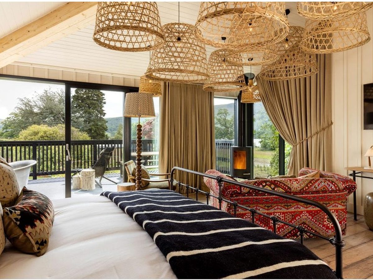 A bedroom with an iron bed, a cluster of woven rattan pendants and a log burner, sliding doors opening to a deck among the trees.