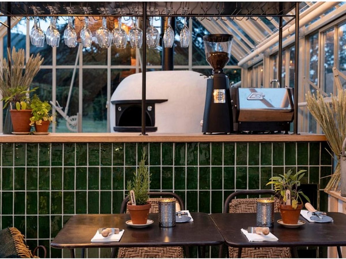 A green-tiled bar with a domed wood-fired pizza oven, timber tables and dried grasses in a glazed room.