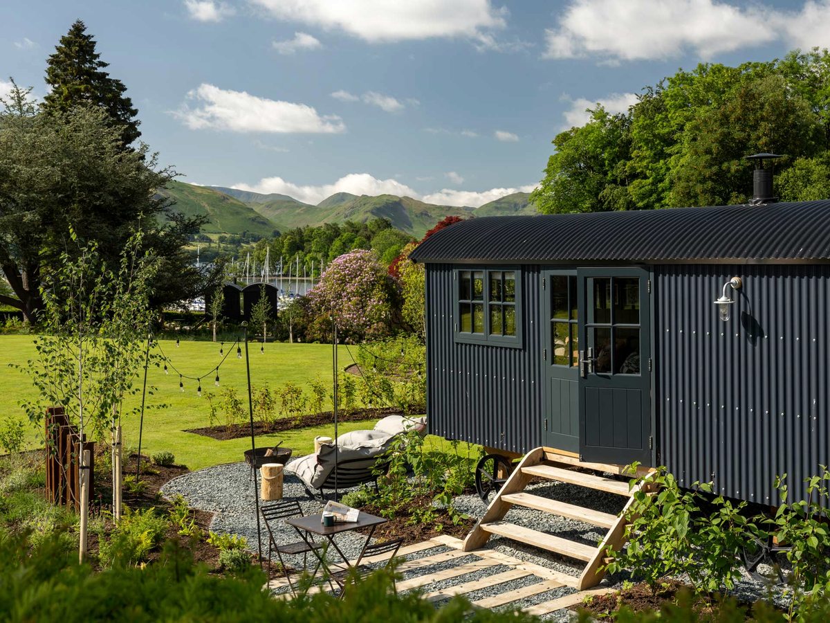 A dark timber shepherd hut with a decked veranda and steps down to a gravel garden with young planting, fells and blue sky behind.