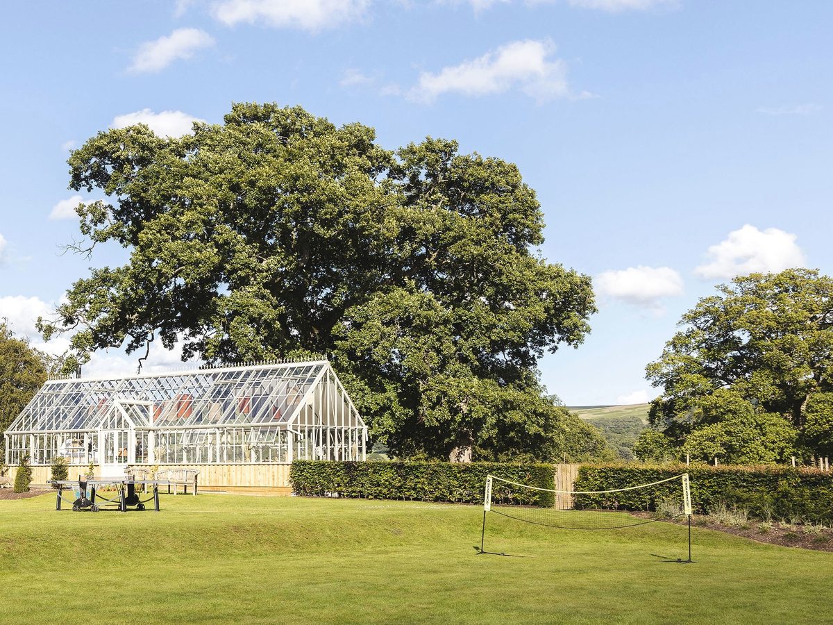 A wide mown lawn with a badminton net in front of a white Victorian glasshouse and a large oak tree, fells beyond under a blue sky.