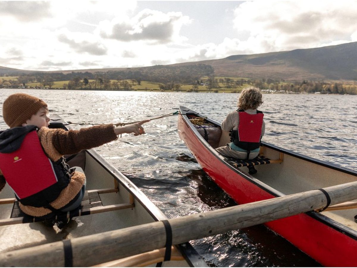 Two open canoes lashed side by side with two children aboard in buoyancy aids, one pointing across the lake.