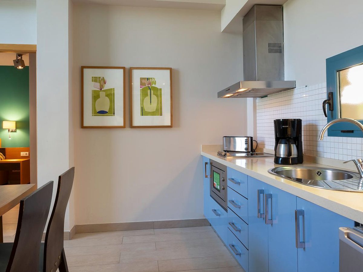 A blue-fronted kitchen with a ceramic hob, oven, extractor hood and coffee maker beside a small dining table.