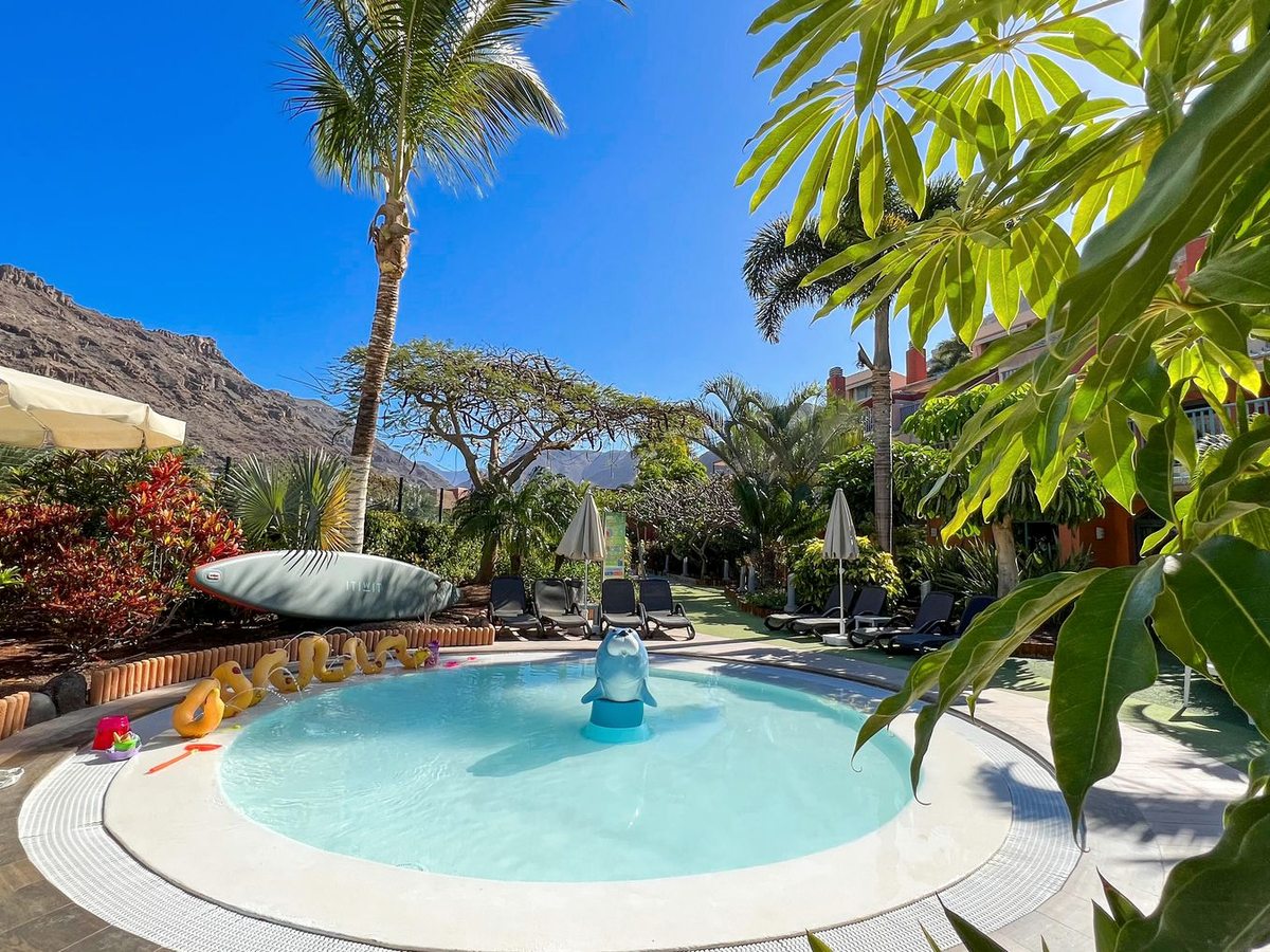A round, shallow children's pool with a dolphin fountain and float toys, palms and mountains behind.
