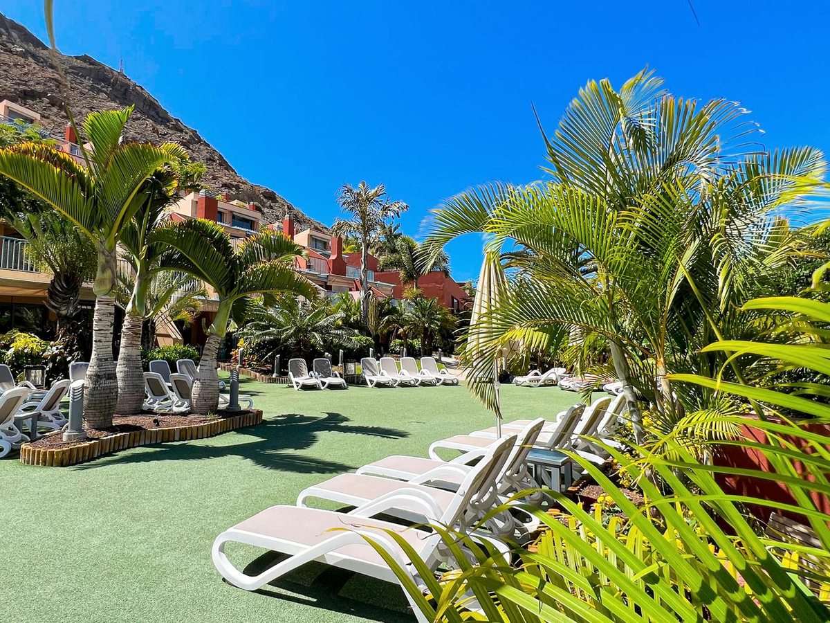 A lawn of white sun loungers among palms and flower beds, with the apartment terraces and mountains rising behind.
