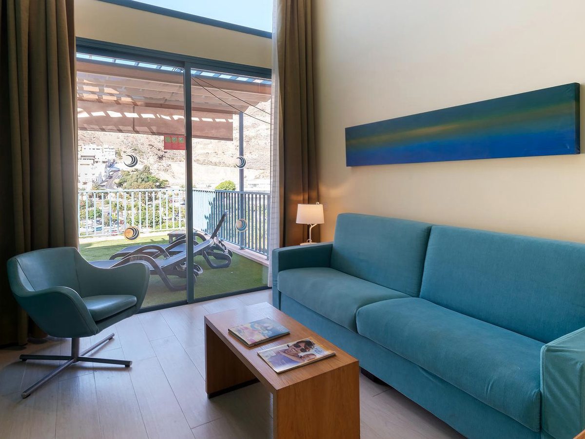 A blue sofa and teal armchair around a wooden coffee table, with sliding doors onto a terrace and a mountain view.