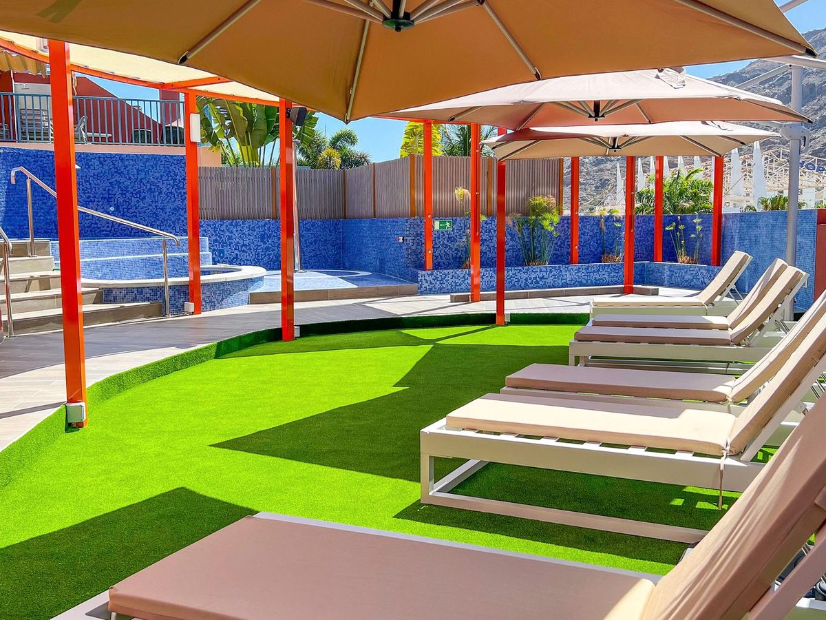 Cream sun loungers under beige parasols on orange poles, on a green artificial-grass lawn beside a blue mosaic-tiled pool, with mountains behind.