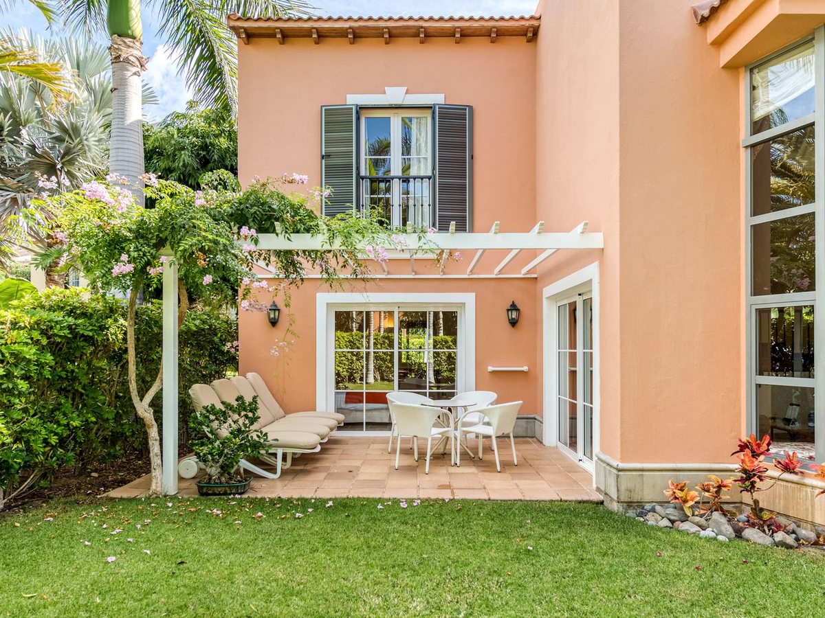 A salmon-pink apartment block with a ground-floor terrace of table and chairs and a cushioned bench under a white pergola of pink bougainvillea, beside a lawn and palms.