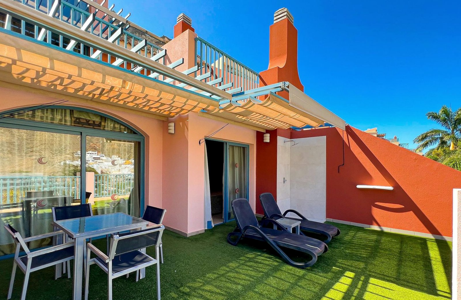 A private apartment terrace at Apartamentos Cordial Mogán Valle, shaded by a pergola awning, with sun loungers and a view over Puerto de Mogán.