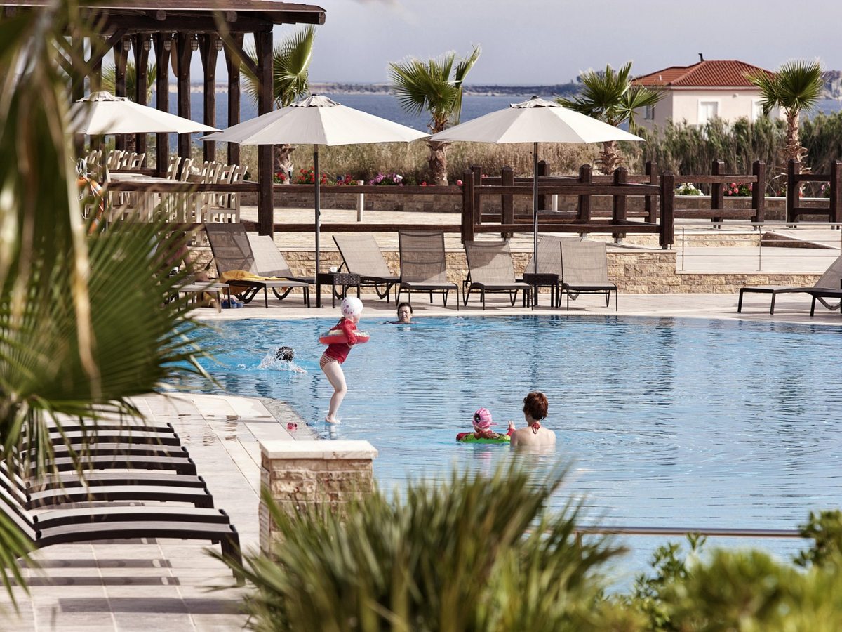 Children and adults swimming in a freshwater pool ringed by loungers, a timber pergola and palms, with the sea beyond.